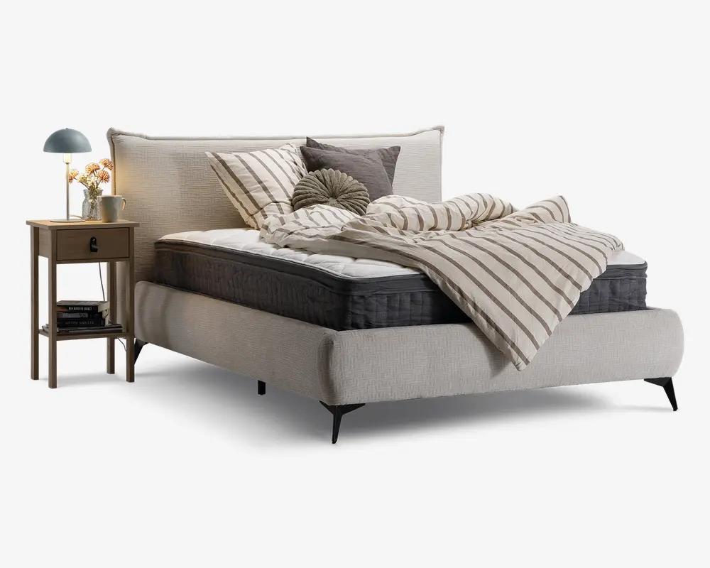 En moderne seng med et lyst beige polstret stel og hovedgærde, udstyret med en tyk, mørkegrå madras. Sengen er redt med en creme- og gråstribet dyne, flere pyntepuder i forskellige former og neutrale nuancer samt et matchende lagen. Ved siden af sengen står et natbord i træ med en skuffe og en hylde, hvorpå der står en blå lampe, en lille vase med blomster og en metal kop. Udtrykket er hyggeligt og moderne.