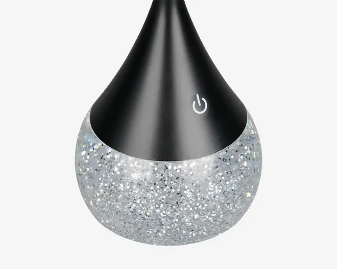 Produktet har et dråbeformet design med en mat sort øverste del og en glitrende, sølvfarvet nederste del. Der er et touchfølsomt tænd/sluk-ikon på den sorte overflade, hvilket indikerer nem betjening. Den glitrende bund giver et dekorativt og stilfuldt udseende, som passer godt til moderne indretning.