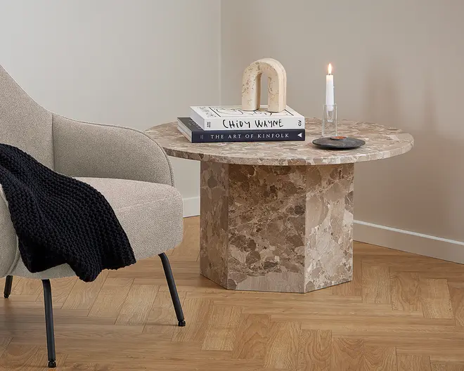 Rundt sidebord lavet af poleret brun marmor med naturlige årer. Bordet har en kraftig, geometrisk piedestalbase og en bred, flad bordplade. På bordpladen ligger to stablede hardback-bøger, en dekorativ bueformet stenfigur og et tændt hvidt stagelys i en klar stage på en mørk coaster. Opsætningen inkluderer en beige polstret lænestol med sorte metalben og et mørkt strikket tæppe lagt over sædet. Gulvet er lyst træ, og væggen er malet i en blød, neutral farve.