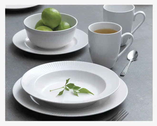Et sæt hvidt porcelænsservice med en diskret rillet kant. Sættet består af en middagstallerken, en dyb tallerken, en skål og et krus. Skålen er fyldt med grønne limefrugter, og kruset indeholder en varm drik. Servicet har et moderne, minimalistisk design med rene linjer og blank finish. En grøn kvist ligger som pynt på den dybe tallerken. Der ses også bestik i rustfrit stål.