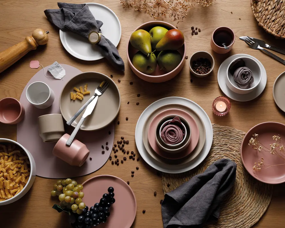 Et koordineret stel bestående af tallerkener, skåle og krus i afdæmpede nuancer af rosa, beige og creme. Sættet inkluderer både runde og ovale tallerkener, stabelbare skåle og matchende krus. Stellet er vist med bestik i rustfrit stål, stofservietter og er stylet med frisk frugt, pasta og kaffebønner for et moderne, rustikt udtryk. Overfladerne har en mat finish, og delene er arrangeret på et træbord med flettede dækkeservietter og dekorative tørrede blomster.