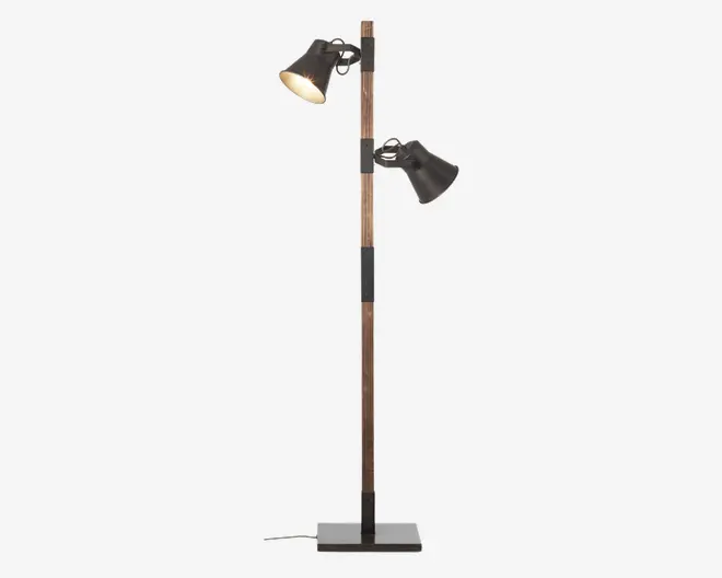 Høj gulvlampe med en slank træstang og to justerbare sorte metalskærme. Hver skærm er fastgjort til stangen med en metalbeslag, så lyset kan rettes i forskellige retninger. Basen er rektangulær og solid, lavet af sort metal. Designet kombinerer moderne og industrielle elementer.