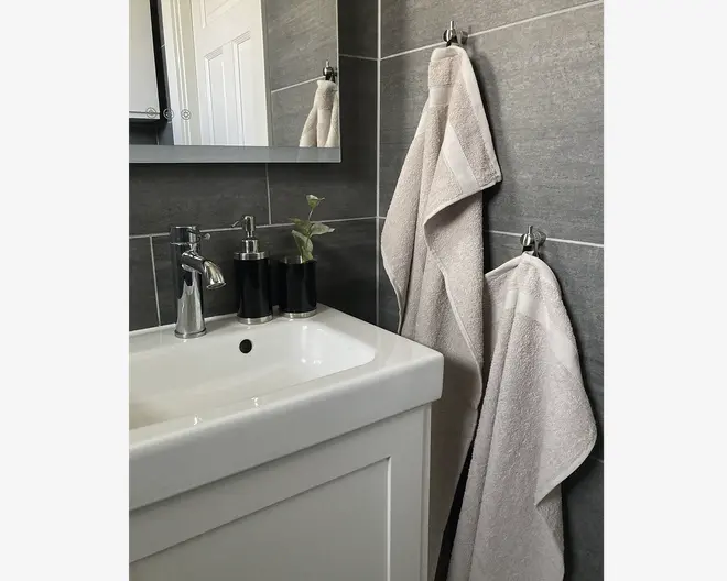 To lysegrå håndklæder med en diskret kantdetalje hænger på kromkroge mod en mørkegrå flisevæg på badeværelset. Håndklæderne ser bløde og fyldige ud med en pænt afsluttet kant. Det større håndklæde hænger øverst, mens det mindre hænger nedenunder. Håndklæderne passer godt til det moderne, minimalistiske badeværelsesdesign.