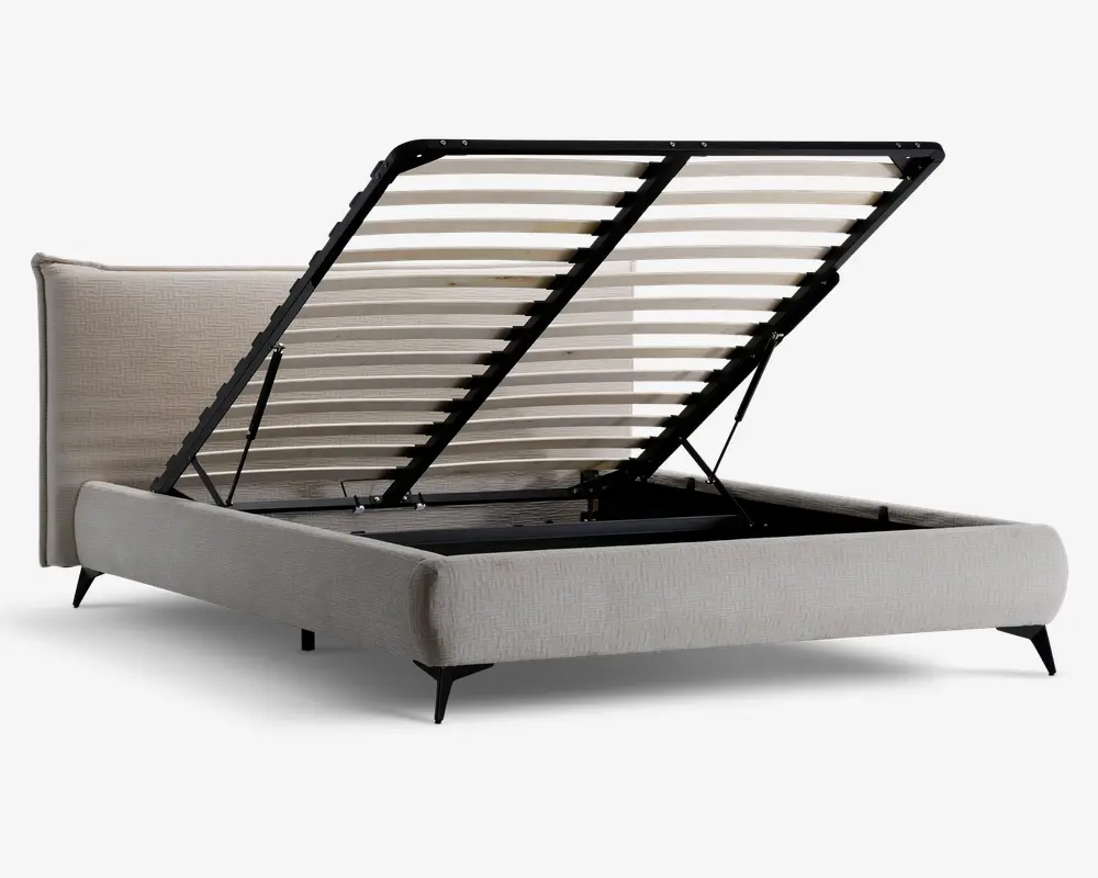 Polstret sengegavl i lysebeige stof med en høj, blød hovedgavl. Lamelbunden er løftet op og afslører et rummeligt opbevaringsrum under sengen. Løftemekanismen understøttes af solide metalarme, som gør det nemt at få adgang til opbevaringsområdet. Sengen står på korte, skrå ben for et moderne udtryk.