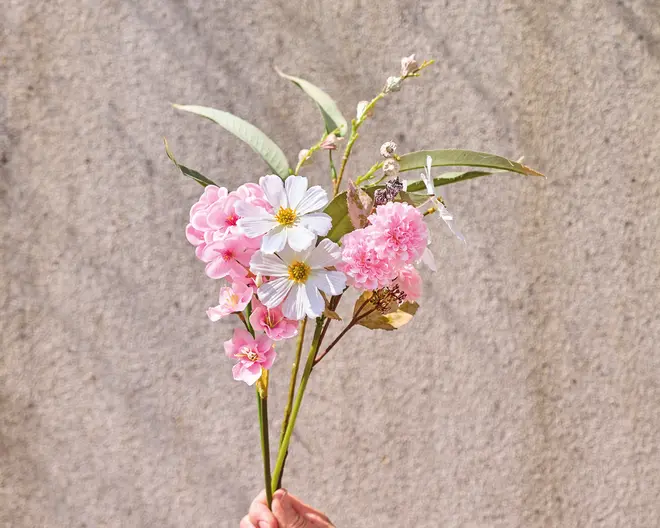 En hånd holder en lille buket med kunstige blomster i lyserøde og hvide nuancer. Buketten indeholder fine blomster med gule midter, fyldige lyserøde blomster og en blanding af grønne og brune blade. Stilkene er lange og slanke, og arrangementet har et naturligt, nyplukket udseende.