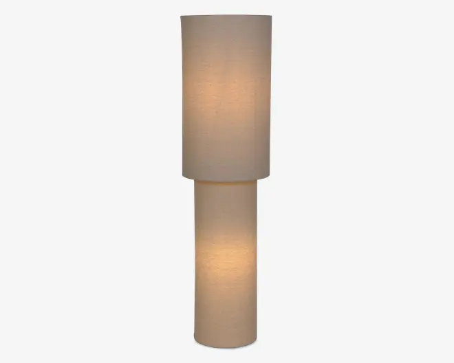 Høj minimalistisk lampe med to stablede cylindriske skærme i beige stof, der lyser blødt med varmt lys.