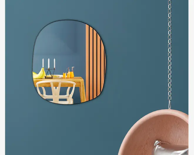 Uregelmæssigt formet vægspejl med en tynd sort ramme, monteret på en mat blå væg. Spejlet reflekterer et moderne spiseområde med et lyst træbord, en hvid stol med buet ryglæn, to hvide lys i en sort stage, en glaskande med appelsinjuice, to glas og en tallerken med bananer. Til højre hænger en stol i lyst træ med en sølvfarvet pude, ophængt i en metal kæde.