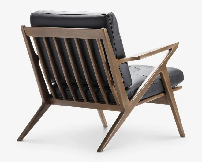 Billedet viser en stol med en moderne, vinklet træramme, der har en slatted ryg og armlæn, som strækker sig i et geometrisk, zigzag mønster. Sæde- og ryghynderne er betrukket med glat, mørkt læder, hvilket giver et elegant og komfortabelt udtryk. Træet har en naturlig finish, der fremhæver dets åre og struktur. Det samlede design kombinerer retrostil med moderne elementer.