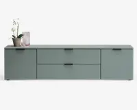 Rektangulært skab med mat salviegrøn finish, to store skuffer i midten og en låge på hver side. Skabet har minimalistiske sorte greb og en glat, flad topplade. Dekorative genstande, herunder en orkidé i potte og en rillet glasvase, står på venstre side af toppen.