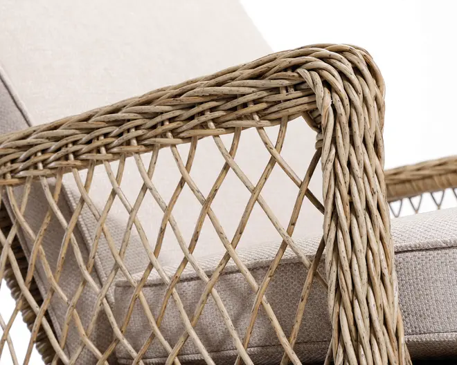 Nærbillede af et armlæn med flettet rattan- eller fletdesign i et krydsmønster. Armlænet er tykt og afrundet med tydelig tekstur fra de naturlige fibre. Sædet og ryglænet er polstret med et lyst beige stof, som giver en blød kontrast til den teksturerede, naturfarvede ramme.