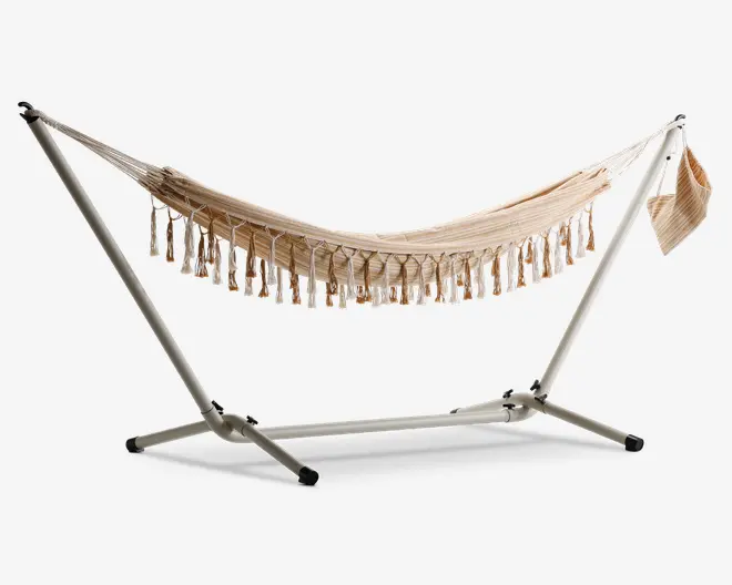 En beige stofhængekøje med dekorative frynser langs siderne, ophængt på et robust, sølvfarvet metalstativ. Hængekøjen har et diskret stribet mønster og en matchende stofpose fastgjort i den ene ende til opbevaring eller tilbehør. Stativet har brede, stabile ben med sorte endestykker for at beskytte gulvet.