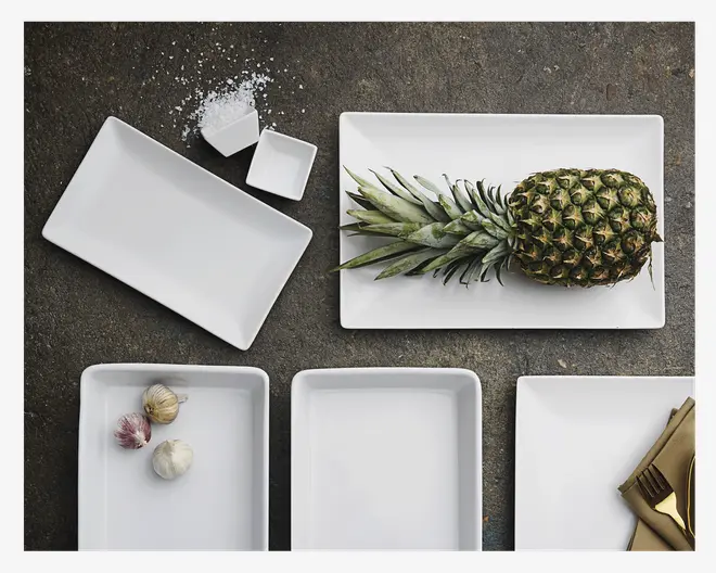 Et sæt hvide rektangulære og firkantede keramiske serveringsfade vist på en mørk overflade. Fadene varierer i størrelse, nogle er dybe og andre lave. Ét fad indeholder en hel ananas, et andet har hvidløg, og to små firkantede fade indeholder salt. Fadene har rene, moderne linjer og en blank finish. Guldfarvet bestik ligger på et af fadene.