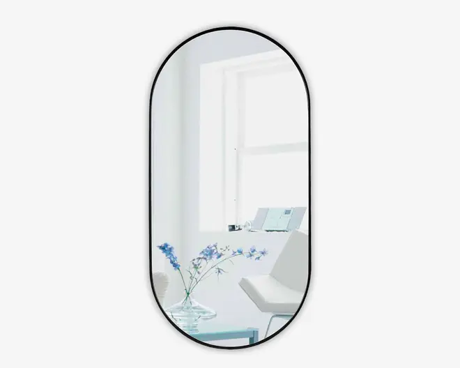 Højt, ovalt spejl med en tynd sort ramme. Spejlet reflekterer et lyst, moderne rum med en hvid stol, en glaskrukke med blå blomster, et lille bord og et stort vindue, der lukker naturligt lys ind. Stilen er minimalistisk og moderne.