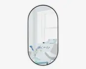 Højt, ovalt spejl med en tynd sort ramme. Spejlet reflekterer et lyst, moderne rum med en hvid stol, en glaskrukke med blå blomster, et lille bord og et stort vindue, der lukker naturligt lys ind. Stilen er minimalistisk og moderne.