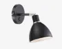Væghængt lampe i mat sort med konisk skærm og rund base. Lampen har en børstet metal-detalje ved samlingen og en indbygget tænd/sluk-knap på basen. Skærmens inderside er hvid for bedre lysrefleksion. Lampens hoved kan justeres for retningsbestemt belysning.