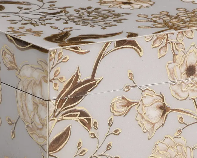 Nærbillede af en æske med cremefarvet baggrund, dekoreret med et indviklet blomstermønster i guld- og brune nuancer. Blomster og blade er fremhævet med metallisk guld, hvilket giver overfladen et let præget og luksuriøst udtryk. Mønstret dækker både låget og siderne, hvilket skaber et sammenhængende og elegant udseende.