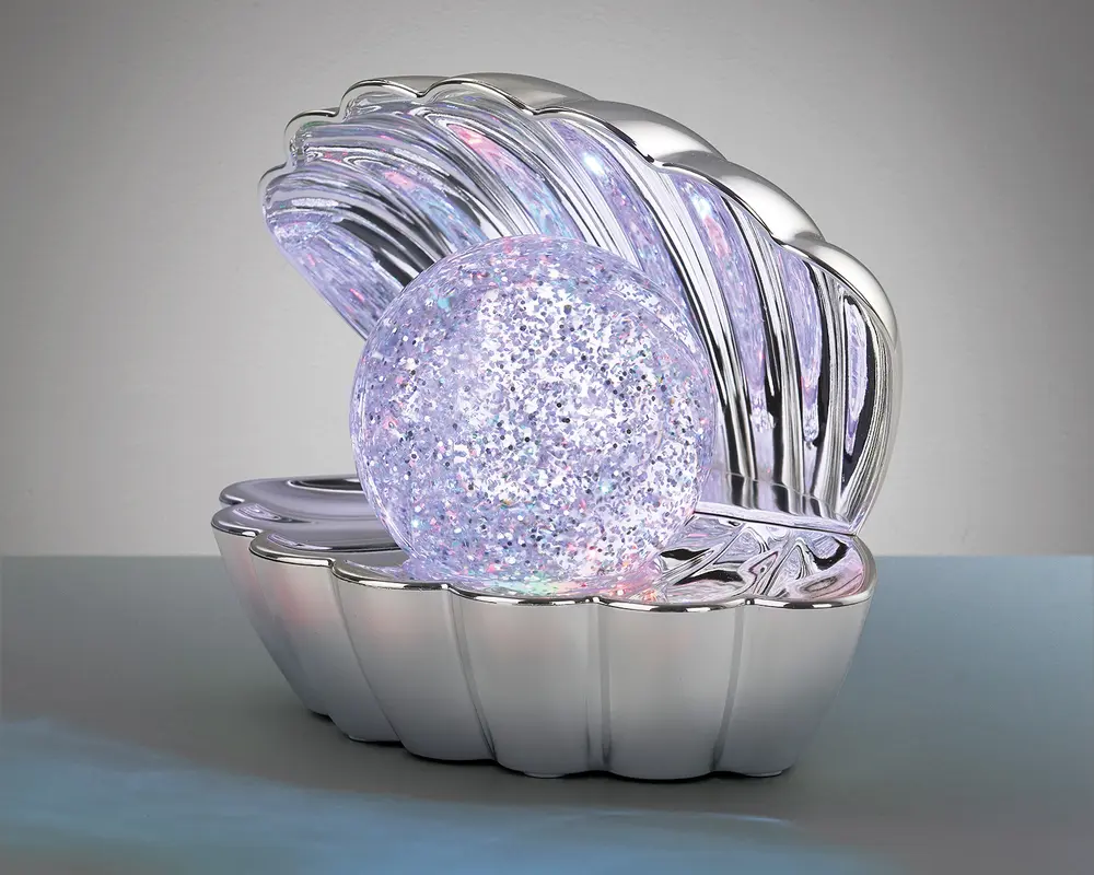 En dekorativ genstand formet som en åben muslingeskal med en blank, sølvfarvet overflade. Inde i skallen ligger en stor, rund kugle fyldt med glimmer, som udsender et blødt, flerfarvet lys. Skallens inderside reflekterer lyset og skaber en glitrende effekt. Det samlede design er elegant og iøjnefaldende, velegnet som natlampe eller pynt.