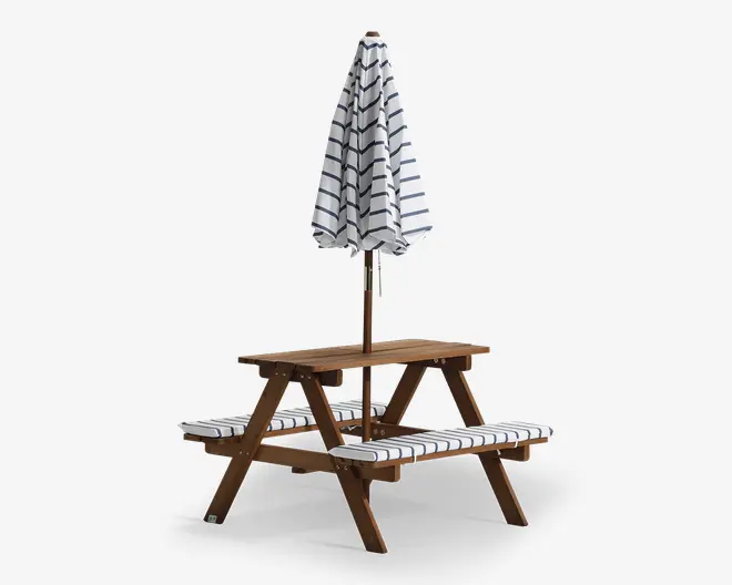 Udendørs bord i træ med to fastmonterede bænke, udstyret med hvide sædehynder med mørkeblå striber. En matchende hvid og mørkeblå stribet parasol er monteret i midten og giver skygge. Bordet og bænkene har et robust, klassisk picnickdesign, velegnet til have eller terrasse.