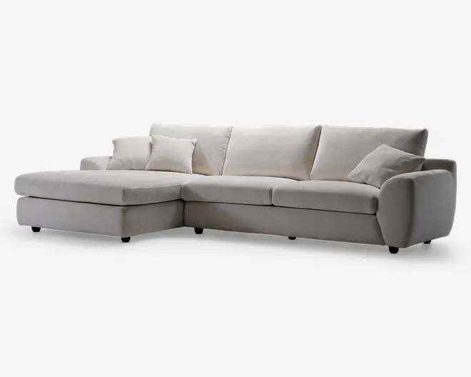 En moderne L-formet sofa med venstrevendt chaiselong, betrukket med lysebeige stof. Sofaen har brede, bløde sædehynder, tre store ryghynder og to matchende pyntepuder. Armlænene er afrundede og polstrede for ekstra komfort, og designet er minimalistisk med rene linjer.