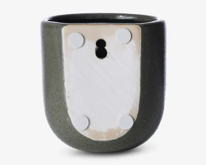 Billedet viser bagsiden af en rund, prikket keramisk beholder med en flad monteringsflade. Monteringsfladen er uglaseret og har fire runde, hvide gummipuder for stabilitet og beskyttelse. I midten er der en nøglehulsformet åbning til ophængning på væg. Resten af beholderen er mørk, prikket og har en glat, blank overflade.