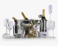 Et festligt arrangement med to flasker champagne i sølv isspande, omgivet af forskellige typer glas, herunder vinglas, champagneglas, cocktailglas og highballglas. Der er også en cocktailshaker i rustfrit stål og flere små diskokugle-dekorationer spredt rundt. Guld- og sølvfarvede serpentiner giver et ekstra festligt præg.
