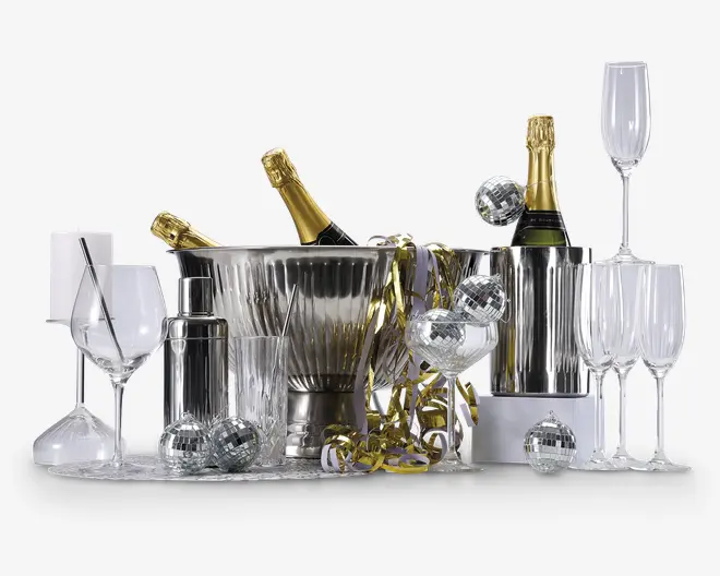 Et festligt arrangement med to flasker champagne i sølv isspande, omgivet af forskellige typer glas, herunder vinglas, champagneglas, cocktailglas og highballglas. Der er også en cocktailshaker i rustfrit stål og flere små diskokugle-dekorationer spredt rundt. Guld- og sølvfarvede serpentiner giver et ekstra festligt præg.