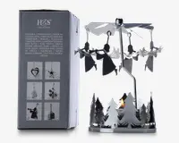 En dekorativ fyrfadsstage i metal med et roterende karruseldesign. Basen er omgivet af udskårne grantræer, og over dem hænger flere engleformede metalfigurer fra en snurrende propel. Når et fyrfadslys tændes i bunden, får varmen propellen og englene til at dreje rundt. Produktet vises ved siden af emballagen, som viser andre tilgængelige designs og produktinformation på flere sprog.