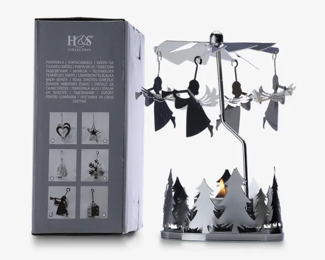 En dekorativ fyrfadsstage i metal med et roterende karruseldesign. Basen er omgivet af udskårne grantræer, og over dem hænger flere engleformede metalfigurer fra en snurrende propel. Når et fyrfadslys tændes i bunden, får varmen propellen og englene til at dreje rundt. Produktet vises ved siden af emballagen, som viser andre tilgængelige designs og produktinformation på flere sprog.