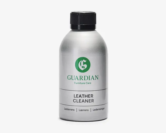 En sølvfarvet flaske med sort skruelåg, der har et grønt logo og mærket 'Guardian Furniture Care' på forsiden. Etiketten angiver tydeligt 'Leather Cleaner' med fed sort skrift. Flasken har et stilrent, moderne design og indeholder produktinformation på flere sprog nederst på etiketten.