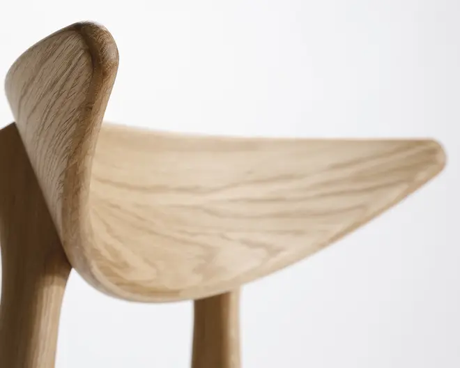 Nærbillede af et træsæde med et glat, buet design og opadgående kanter. Træets årer er tydelige og fremhæver den naturlige struktur og finish. Sædet er ergonomisk formet for komfort med afrundede hjørner og en sømløs konstruktion.