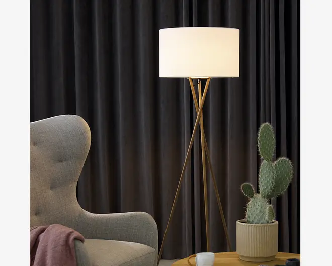 En moderne gulvlampe med en stor, cylindrisk hvid lampeskærm og tre slanke, guldfarvede metalben i et stativdesign. Lampen står ved siden af en lysegrå polstret lænestol med buet ryg og et lyserødt tæppe lagt over armlænet. Der er også en rillet beige keramikpotte med en høj, grøn kaktus med flere puder, placeret på et træsidebord. Baggrunden har mørke, tunge gardiner.