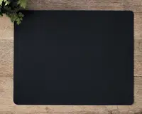 Et rektangulært, ensfarvet sort underlag med afrundede hjørner, placeret på en træoverflade. Underlaget har en glat overflade og et minimalistisk design uden synlige mønstre eller logoer. En lille buket grønne blade ses i øverste venstre hjørne.