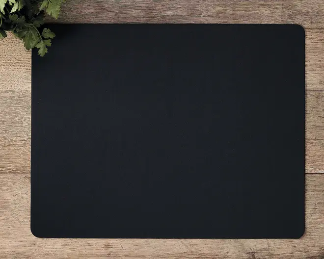 Et rektangulært, ensfarvet sort underlag med afrundede hjørner, placeret på en træoverflade. Underlaget har en glat overflade og et minimalistisk design uden synlige mønstre eller logoer. En lille buket grønne blade ses i øverste venstre hjørne.