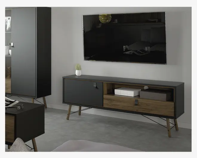 Et moderne TV-bord med mat sort finish og trædetaljer. Møblet har skråtstillede træben, en stor sort låge til venstre og to åbne hylder til højre – den ene med træfront og lille greb. På den åbne hylde står en hvid æske og en grå æske. Bordpladen er dekoreret med en lille potteplante. Designet er minimalistisk og nutidigt.