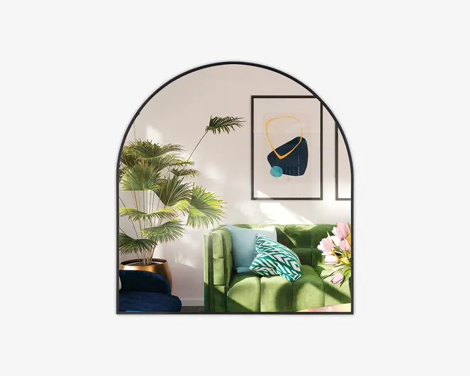 Et moderne stueinteriør med en blød grøn veloursofa, der har to pyntepuder – én med et geometrisk blåt og hvidt mønster og én i ensfarvet lyseblå. Sofaen er suppleret med en buket lyserøde liljer på et sidebord. Bag sofaen står en stor pottepalme, som tilfører rummet grønne nuancer. Væggen er dekoreret med to abstrakte indrammede kunstværker, og rummet er oplyst af naturligt sollys, hvilket skaber en lys og indbydende stemning.