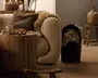 Hyggelig stue i neutrale farver med en buet beige sofa, en kraftig brun teksturpude, et pelslignende tæppe, et rundt sidebord i træ med en kop og stearinlys samt en sort kurv med foldede tæpper ved siden af en lille træskammel og vase.