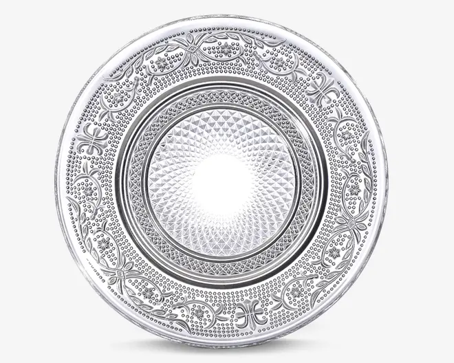 Rund tallerken med indviklet præget mønster. Midten har et geometrisk diamantmønster, omgivet af et bånd med små hævede prikker. Den ydre kant er dekoreret med elegante blomster- og vinstokmotiver, hvilket giver tallerkenen et dekorativt og elegant udseende. Overfladen har en blank, reflekterende finish.