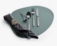 Et sæt med fire bestikdele i rustfrit stål, herunder en kniv, gaffel, spiseske og teske, alle med et stilrent, moderne design og blank finish. Bestikket er pænt arrangeret på en mat, uregelmæssigt formet dækkeserviet. En mørkegrå stofserviet er foldet og holdt sammen af en rund servietring i guldfarve med marmorlignende mønster.