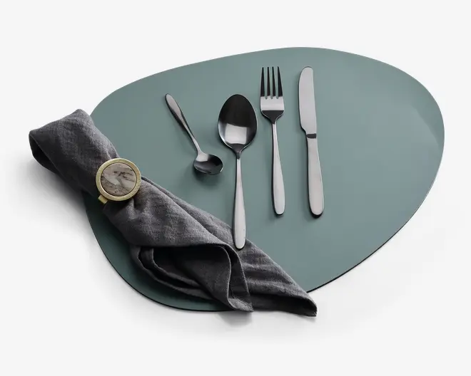 Et sæt med fire bestikdele i rustfrit stål, herunder en kniv, gaffel, spiseske og teske, alle med et stilrent, moderne design og blank finish. Bestikket er pænt arrangeret på en mat, uregelmæssigt formet dækkeserviet. En mørkegrå stofserviet er foldet og holdt sammen af en rund servietring i guldfarve med marmorlignende mønster.