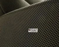 Nærbillede af en tekstureret stofoverflade i en mørk, neutral farve. Stoffet har et hævet, bikube-lignende mønster, som giver en taktil og visuelt interessant finish. Et lille rektangulært mærke med teksten 'VIOLINO' er syet på stoffet og angiver producenten. Syningen langs kanten er synlig, hvilket tyder på holdbarhed og omhu i konstruktionen.