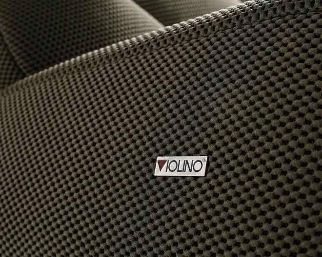 Nærbillede af en tekstureret stofoverflade i en mørk, neutral farve. Stoffet har et hævet, bikube-lignende mønster, som giver en taktil og visuelt interessant finish. Et lille rektangulært mærke med teksten 'VIOLINO' er syet på stoffet og angiver producenten. Syningen langs kanten er synlig, hvilket tyder på holdbarhed og omhu i konstruktionen.