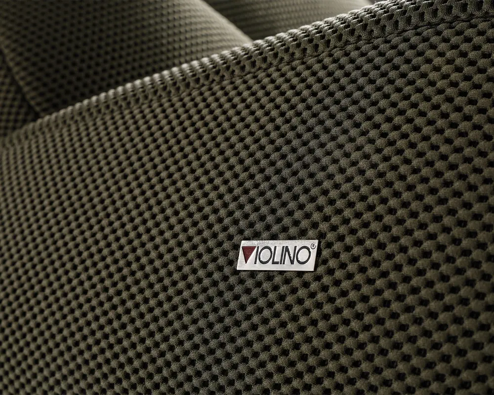 Nærbillede af en tekstureret stofoverflade i en mørk, neutral farve. Stoffet har et hævet, bikube-lignende mønster, som giver en taktil og visuelt interessant finish. Et lille rektangulært mærke med teksten 'VIOLINO' er syet på stoffet og angiver producenten. Syningen langs kanten er synlig, hvilket tyder på holdbarhed og omhu i konstruktionen.