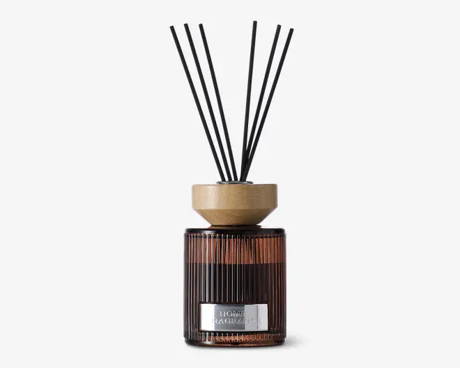 En dekorativ glasbeholder med lodret rillet struktur og mørk ravfarvet tone. Den har et lyst trælåg og flere tynde sorte duftpinde, der stikker op fra toppen. Beholderen har en metallisk label på forsiden, hvilket giver et moderne udtryk.