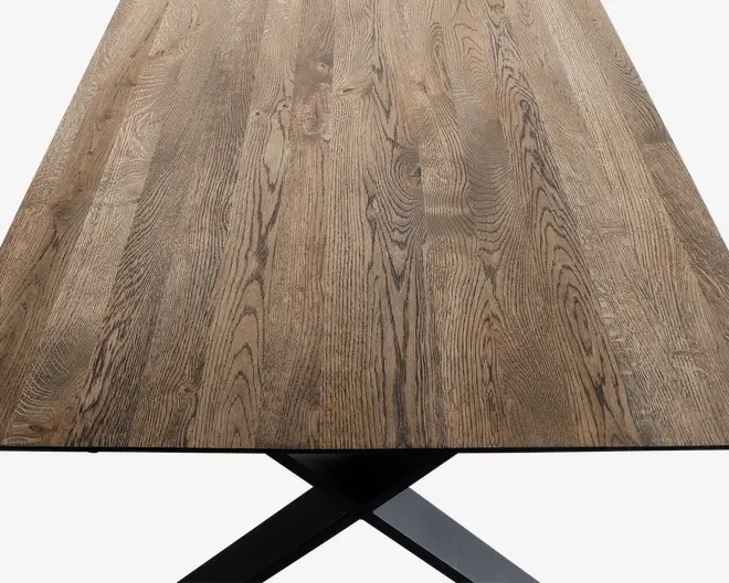 Rektangulær bordplade lavet af naturtræ med synligt åremønster og en glat, mat overflade. Træet har en varm, mellembrun tone med diskrete farvevariationer. Bordet understøttes af en solid, sort metalbase i krydsform, som delvist kan ses nederst på billedet.