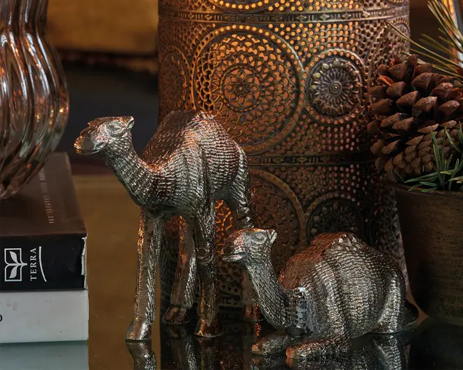 To metalliske kamelfigurer med en rillet, tekstureret overflade. Den ene kamel står op, mens den anden ligger ned. Begge har en skinnende, sølvfarvet finish, der reflekterer lyset. Figurerne står på en reflekterende overflade ved siden af en dekorativ, mønstret metalkrukke og en lille dekoration med kogler og grønt.