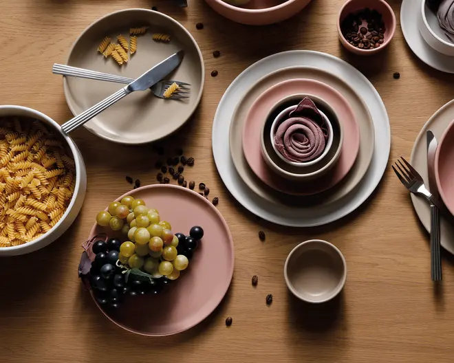 Et sæt keramikservice i afdæmpede rosa og beige nuancer, herunder tallerkener, skåle og kopper arrangeret på en træoverflade. Sættet har glatte, matte overflader og et minimalistisk design. Nogle skåle er placeret inde i større tallerkener, og sættet suppleres af bestik i rustfrit stål. Billedet viser også madvarer som vindruer, pasta og kaffebønner, hvilket fremhæver servicets alsidighed til servering af forskellige typer mad.