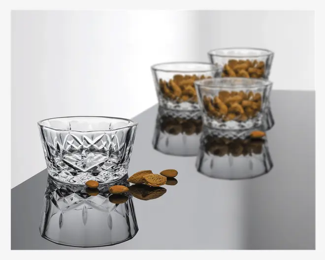 Sæt med fire klare glasskåle med facetslebet, diamantmønstret design på sider og bund, hvilket giver dem et glitrende og elegant udseende. Skålene har en bred åbning og en solid, tyk bund. Tre af skålene er fyldt med mandler, mens én skål er tom, hvilket fremhæver det detaljerede mønster. Den reflekterende overflade under skålene forstærker deres visuelle udtryk.