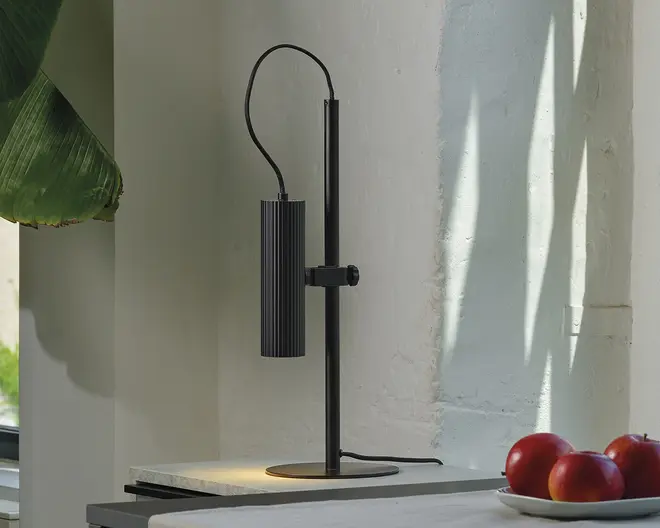 En moderne, minimalistisk bordlampe med mat sort finish. Lampen har en lodret cylindrisk skærm med rillet tekstur, ophængt fra en justerbar arm, der er fastgjort til en rund fod. Strømkablet er synligt og buer fra toppen af lampen. Lampen står på en lys overflade tæt ved en væg, hvor naturligt lys kaster skygger i nærheden.