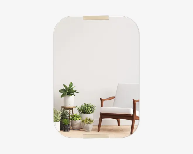 Rektangulært spejl med afrundede hjørner og et tyndt, rammeløst design. Spejlet har to små, guldfarvede monteringsbeslag øverst og nederst. Refleksionen viser et moderne interiør med en lænestol i træ med hvide hynder i retrostil samt flere potteplanter i forskellige størrelser og teksturer, placeret på gulvet og et lille sidebord.