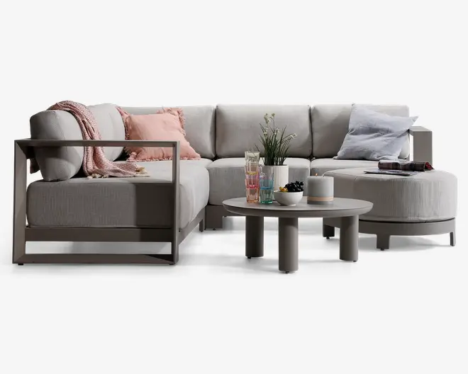 Moderne hjørnesofa med lysegråt betræk og armlæn i metalramme. Sofaen har bløde, dybe puder og inkluderer pyntepuder i pastelrosa og lyseblå samt et tekstureret tæppe. Foran sofaen står et rundt sofabord med mat finish, hvor der er en plante, farverige glas, en skål med bær og et stearinlys. Opsætningen giver et indtryk af en komfortabel og stilfuld stue med moderne design.